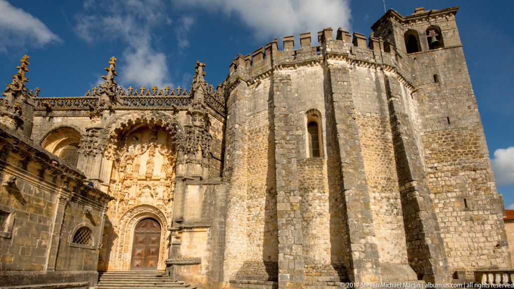 Wide exterior view of the Convent of Christ church with ornate portal, massive polygonal stone apse, crenellated parapet, and square bell tower.