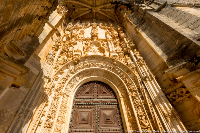 Steep low-angle view of a tall ornate portal with wooden doors beneath layered carved arches, niches, figures, foliage, and vertical shafts.