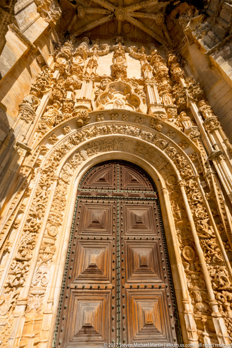 Low-angle view of an ornate church portal with wooden double doors, concentric carved arches, slender colonnettes, figures, and sculptural cresting.