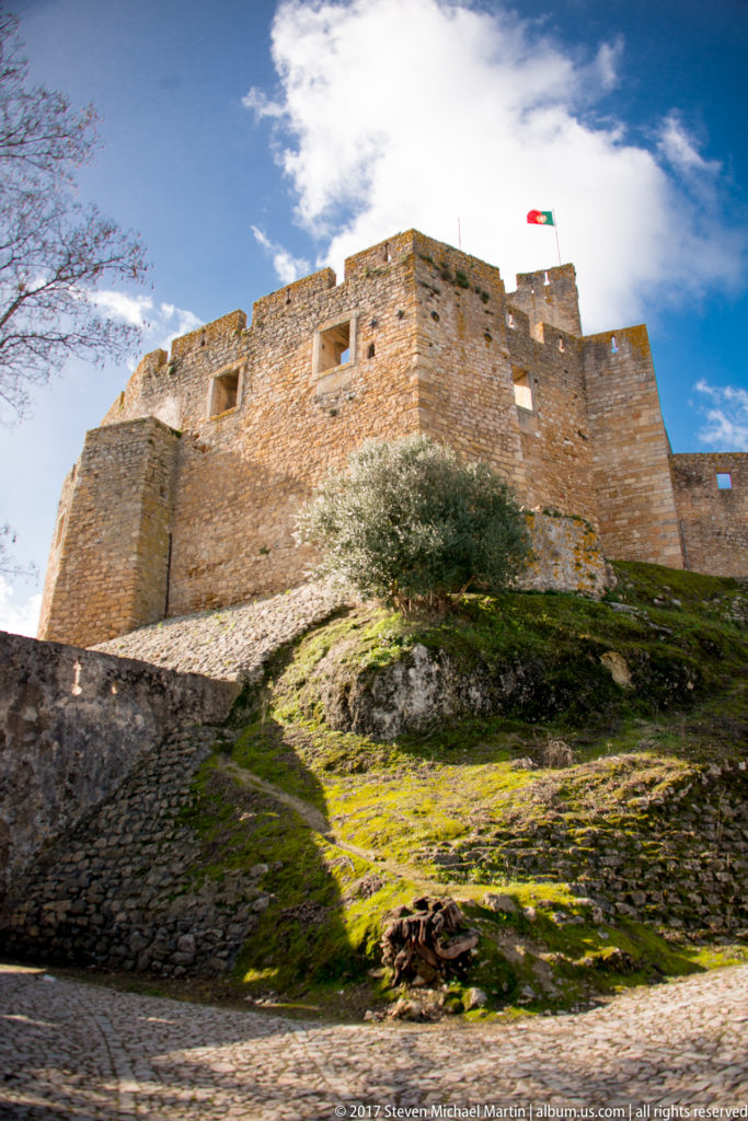 Wide low-angle view of stone castle walls with projecting towers, Portuguese flag, mossy hillside, grass, and an olive tree.