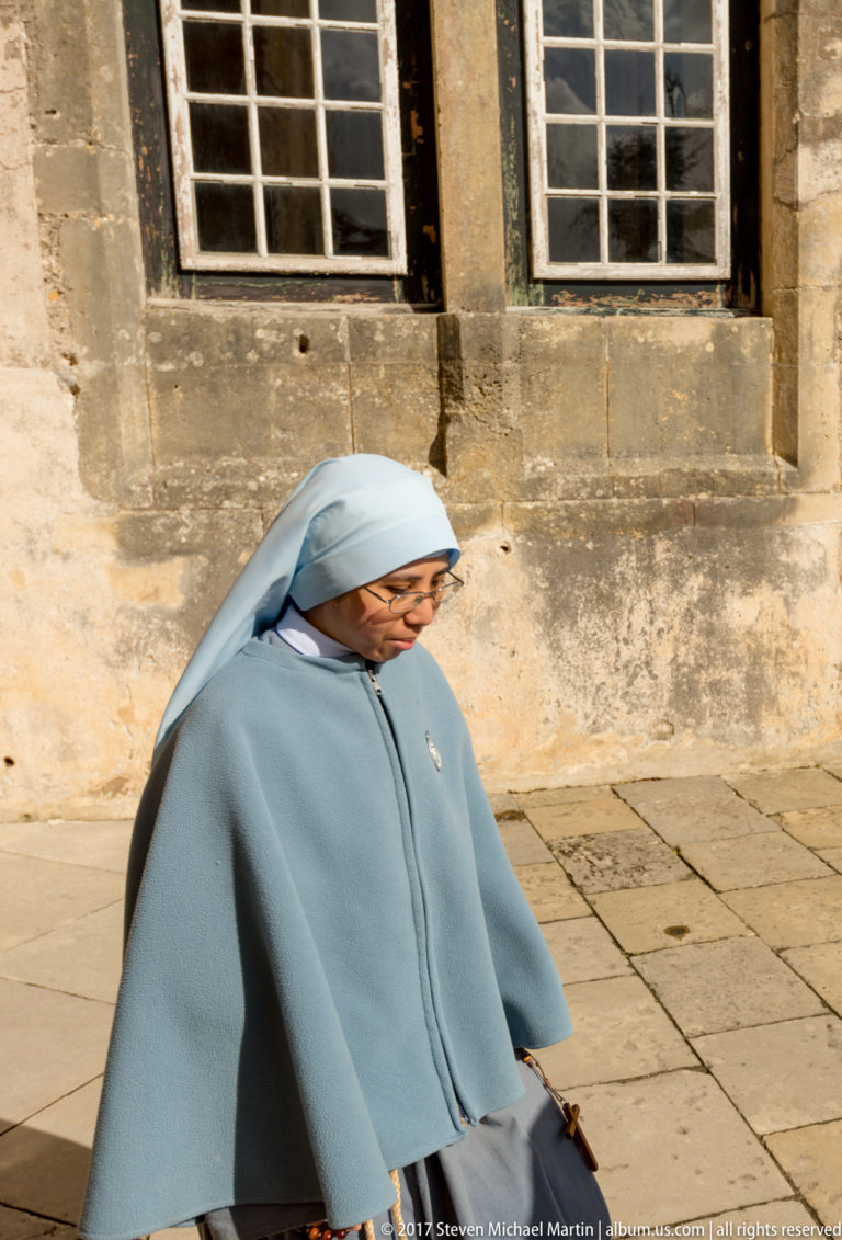 Woman in a pale blue-gray nun's habit and veil walking across sunlit stone paving beside old stone walls and windows.