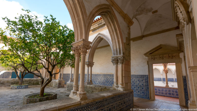 Small cloister courtyard with a citrus tree, pointed arches on slender columns, stone paving, and blue-and-white tiled lower walls.