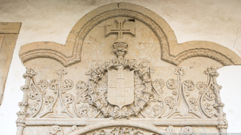 Portuguese heraldic shield surrounded by carved scrollwork and pinnacles, with a cross above inside a rounded stone architectural frame.