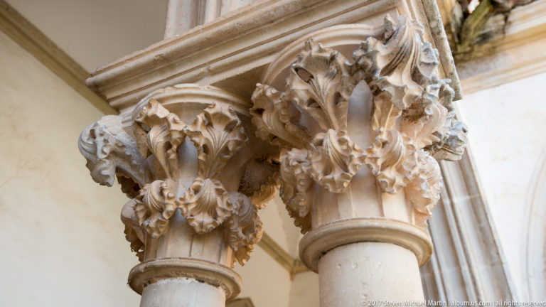 Paired pale stone columns with elaborate leafy capitals beneath molded arches, seen from an oblique upward angle.