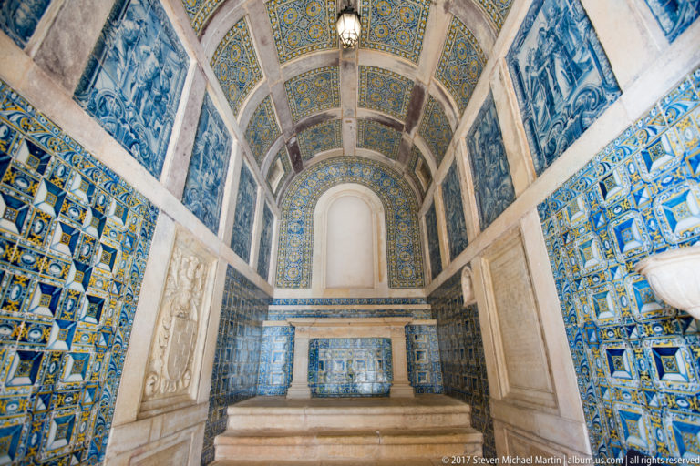 Narrow barrel-vaulted chapel lined with blue-and-yellow patterned tiles and blue-and-white narrative panels, ending at a raised stone altar area.