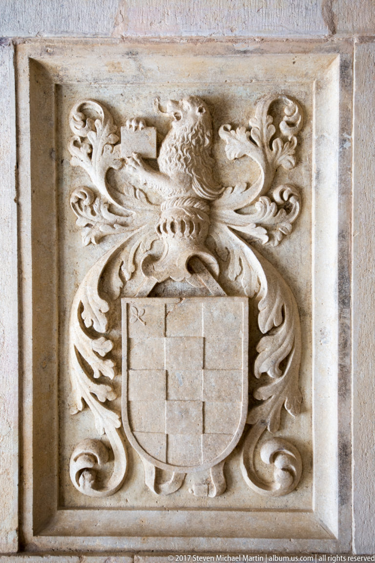 Carved Portuguese shield beneath a crested helmet, surrounded by deeply scrolling heraldic mantling in a rectangular stone panel.