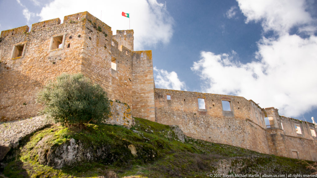 Stone castle walls and tower with Portuguese flag above a grassy rocky slope and olive tree under a bright clouded sky.