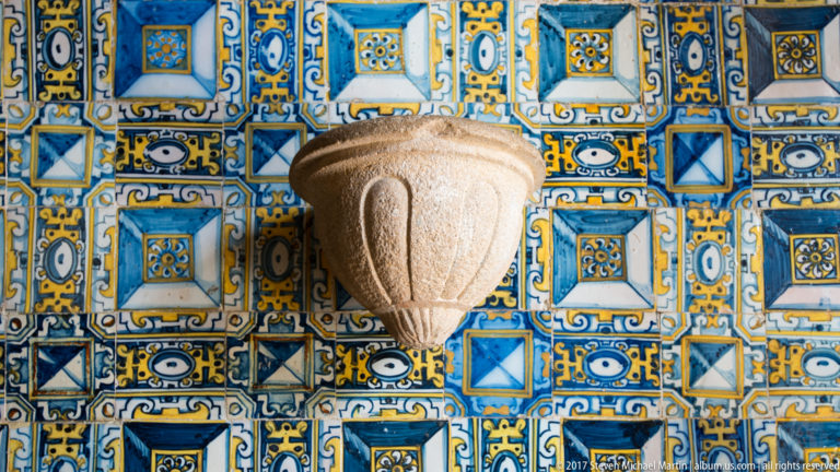 Small carved stone basin projecting from a wall covered in bright blue, yellow, and white patterned ceramic tiles.