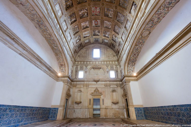 Axial view down a long hall with richly coffered barrel vault, white walls, blue tile dado, high windows, and distant doorway.