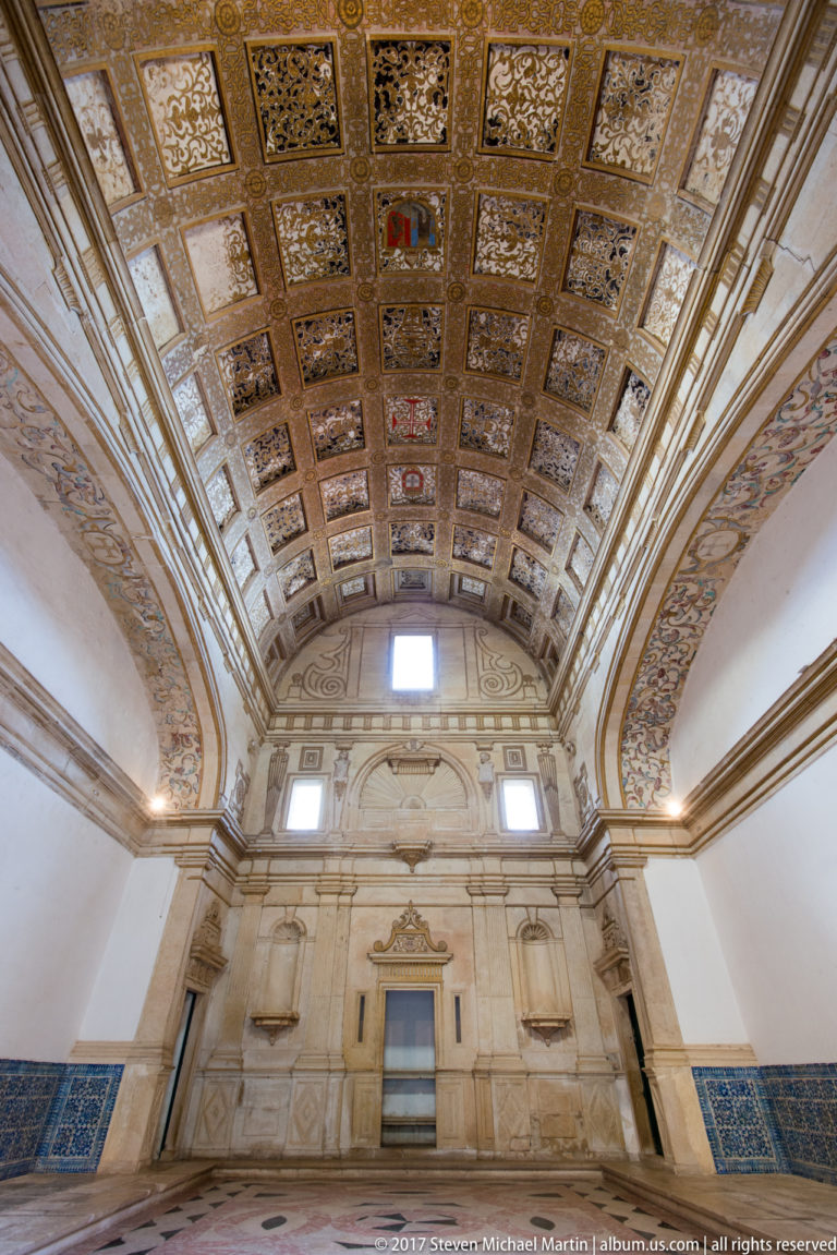 Long pale hall with an ornate coffered barrel ceiling, classical wall niches and doorways, high windows, and blue tile panels.