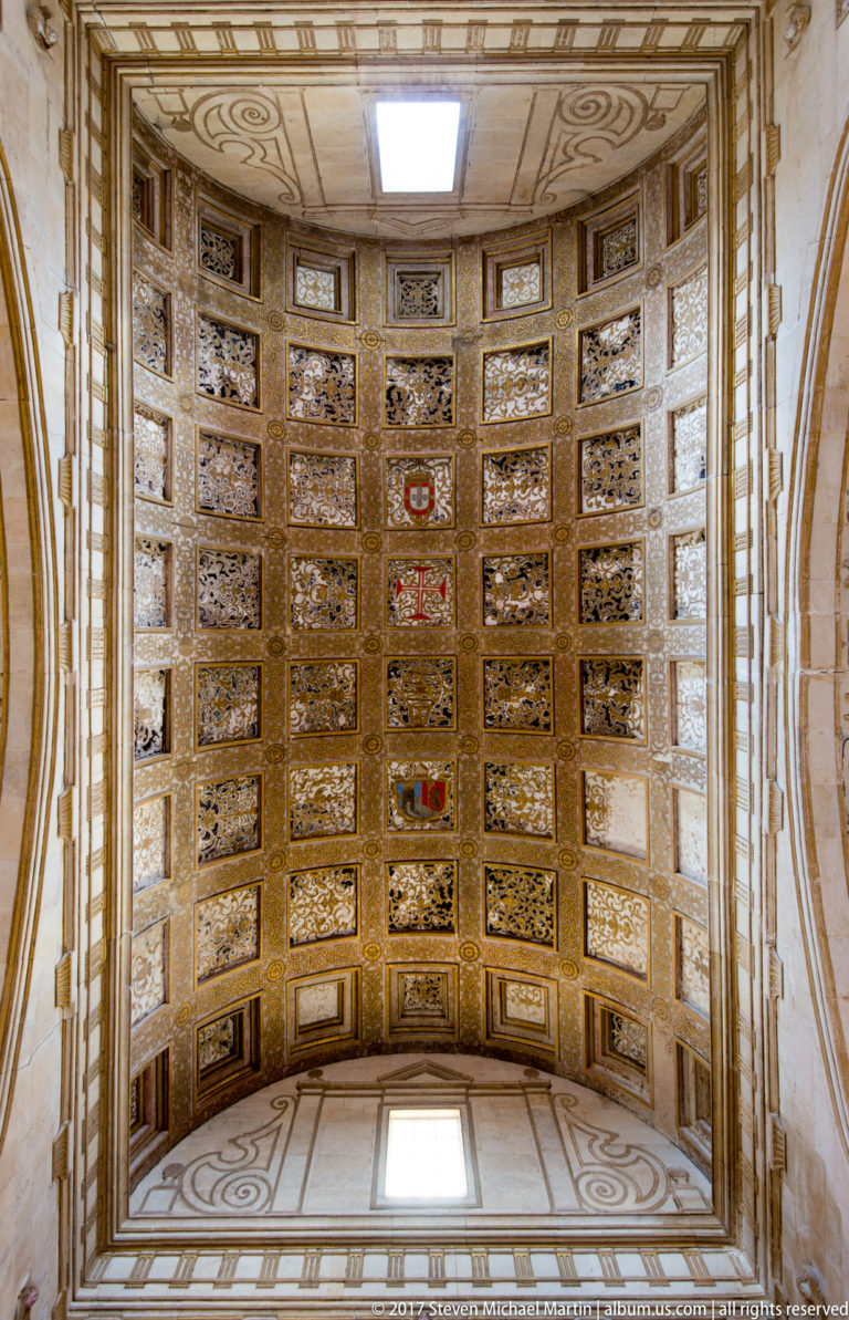 Symmetrical upward view of an ornate barrel ceiling filled with carved and painted square coffers and small red heraldic accents.