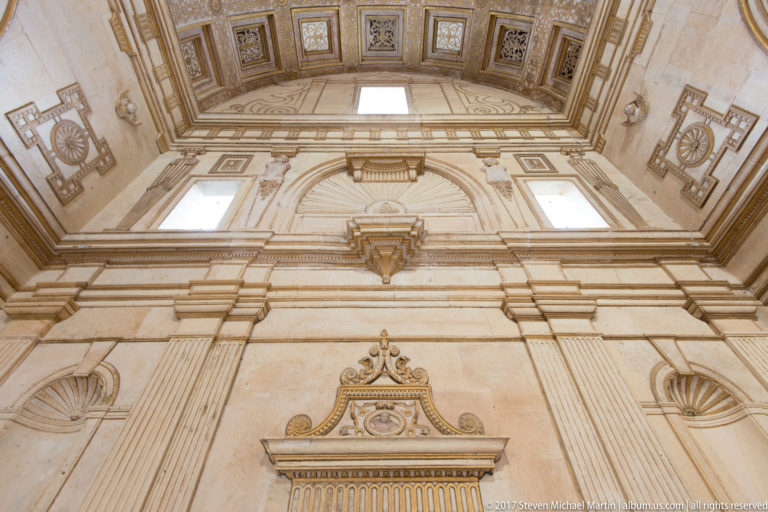 Upward view of a tall pale-stone interior wall with pilasters, niches, rectangular windows, shell motifs, and a decorated coffered ceiling.