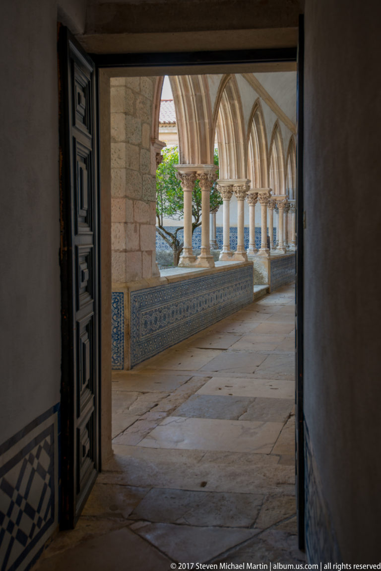 View from a dark doorway into a bright cloister with pointed arches, slender columns, blue tiled walls, stone paving, and a tree.