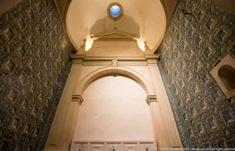 Tall pale stone arched niche between walls covered in blue-and-yellow patterned azulejos, with a small round window above.