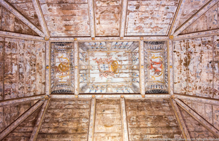 Weathered painted wooden ceiling divided into rectangular coffers with heraldic shields, floral designs, and faded figurative decoration.
