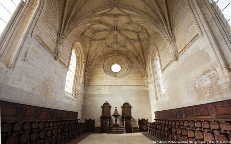 Long stone choir room with wooden stalls on both sides, ribbed vaulting, tall windows, circular end window, and three high-backed seats.
