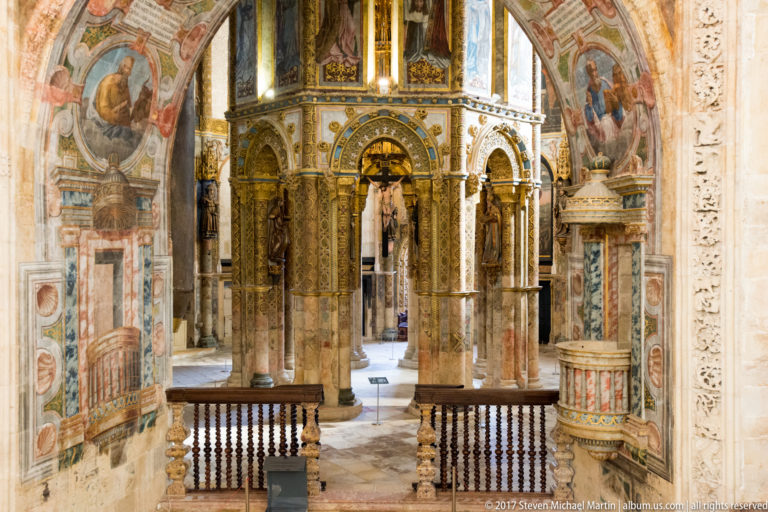 Interior view across a wooden balustrade toward the Charola’s columned central core, surrounded by murals, gilding, and painted vaults.