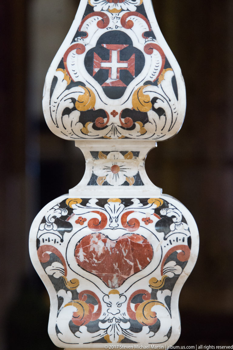 White cross in a dark upper medallion above a red heart-shaped stone inset surrounded by colorful scrolling ornament.