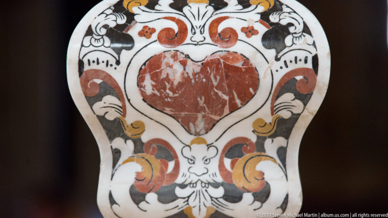 Heart-shaped red stone inset surrounded by symmetrical black, orange, gold, and white scrolling ornament and grotesque faces.