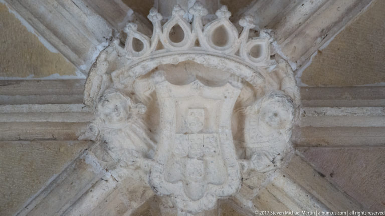 Crowned stone heraldic shield with small flanking figures set at the intersection of diagonal vault ribs.