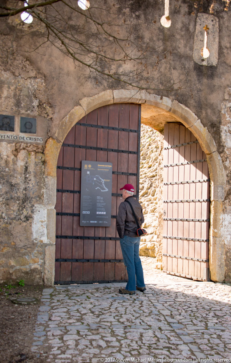 Visitor in a red cap stands before a broad arched stone entrance with partly open timber gates and an interpretive sign.