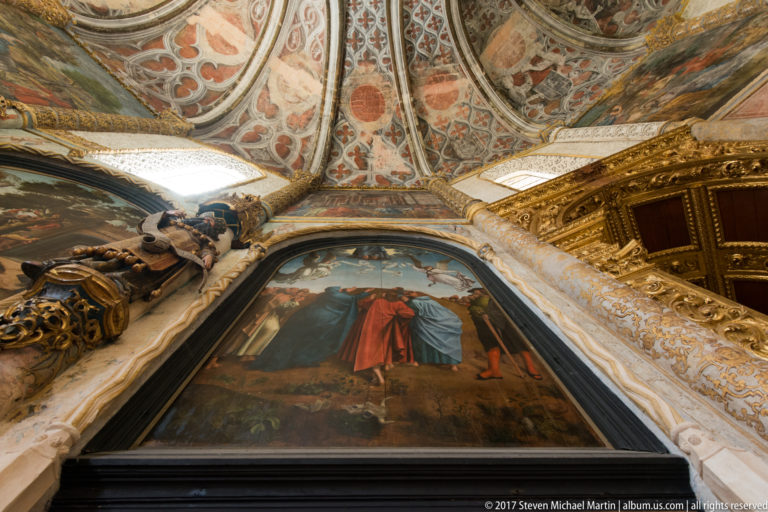 Low-angle interior view with a large religious painting beneath painted vaults, gilded carving, and a projecting sculpted figure.