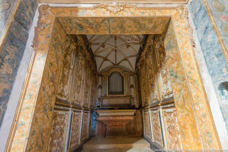 Ornate doorway into a narrow painted chapel-like space with gilded wall panels, blue surfaces, ribbed vaulting, and an altar-like block.