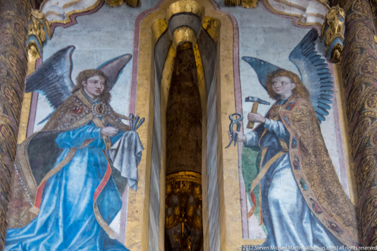 Two painted winged angels in colorful robes flank a narrow vertical opening within an ornate interior wall.