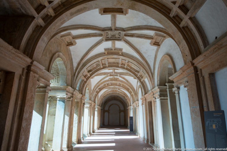 Long rib-vaulted cloister gallery with pale stone columns, white walls, and alternating bands of sunlight along the floor.