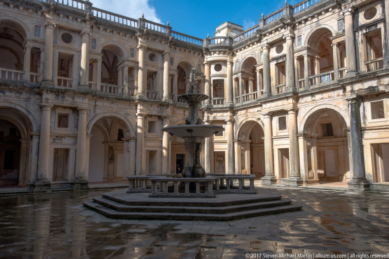 Sunlit oblique view of a two-story Renaissance cloister with central fountain, arched galleries, columns, and balustrades.