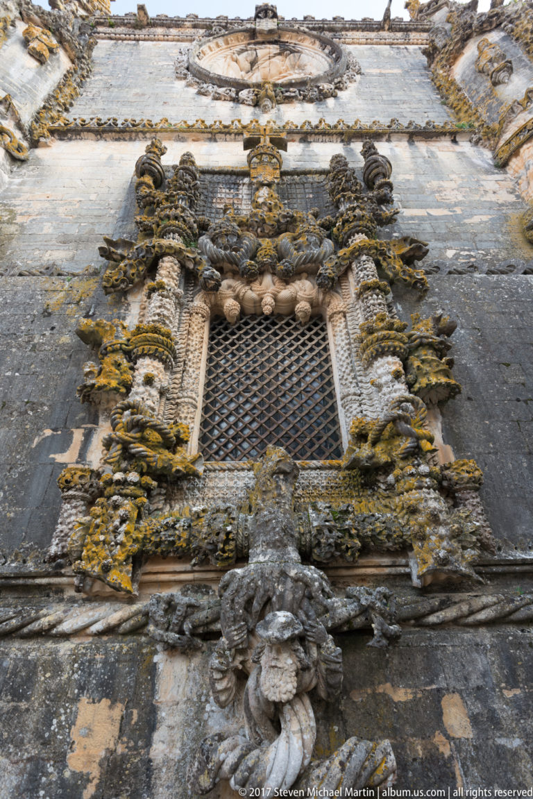 Frontal gridded window with clustered carved shafts, rope-like motifs, heraldic elements, and projecting sculpture on a dark stone wall.