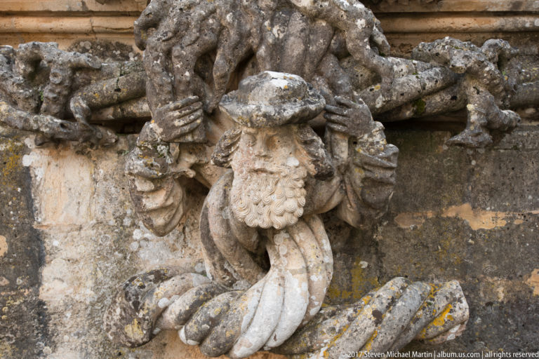 Weathered bearded stone figure with curled beard and raised arms beneath projecting exterior carvings.