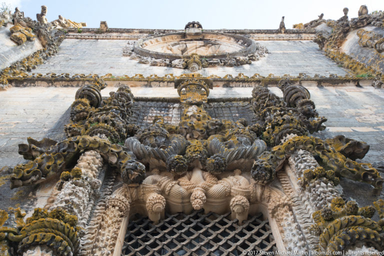 Low-angle view up an ornate gridded window with twisted stone shafts, projecting carvings, heraldic motifs, and a circular feature above.
