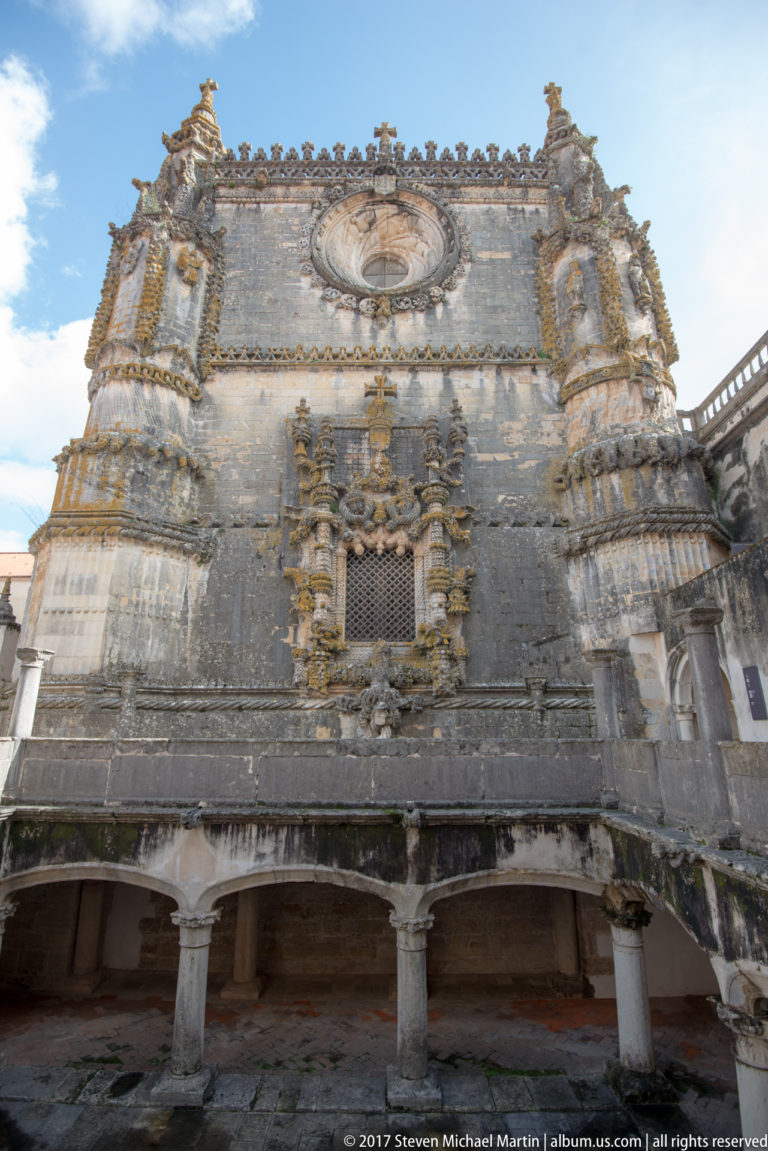Tall stone façade with a large circular window above an ornate gridded opening, framed by buttresses and a ground-level arcade.