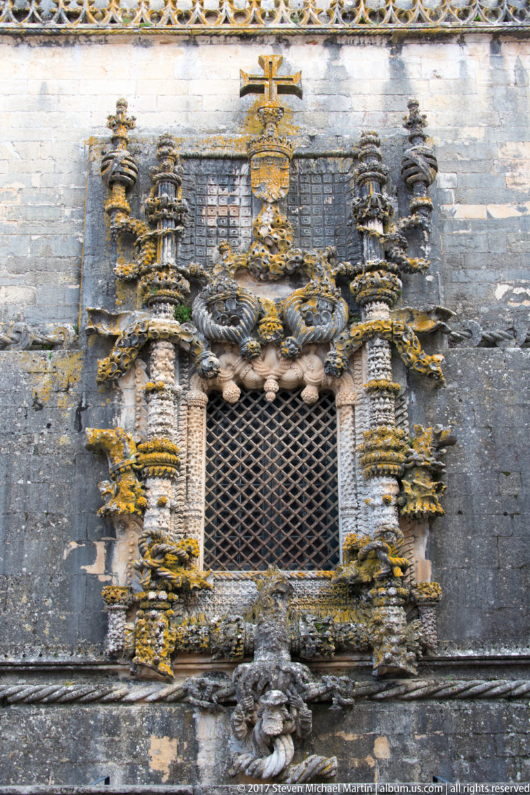 Front-facing gridded window with elaborate projecting stone surround, twisted shafts, heraldic forms, and a cross above.