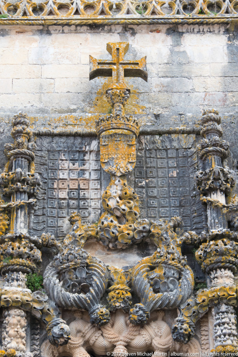 Close-up of a cross and heraldic crown above dense rope-like carved stone ornament on a dark exterior wall.