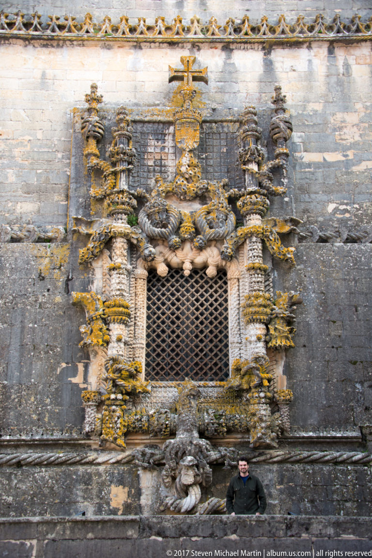 Small gridded window surrounded by elaborate carved stonework, heraldic emblems, twisted shafts, and a cross, with a person below for scale.