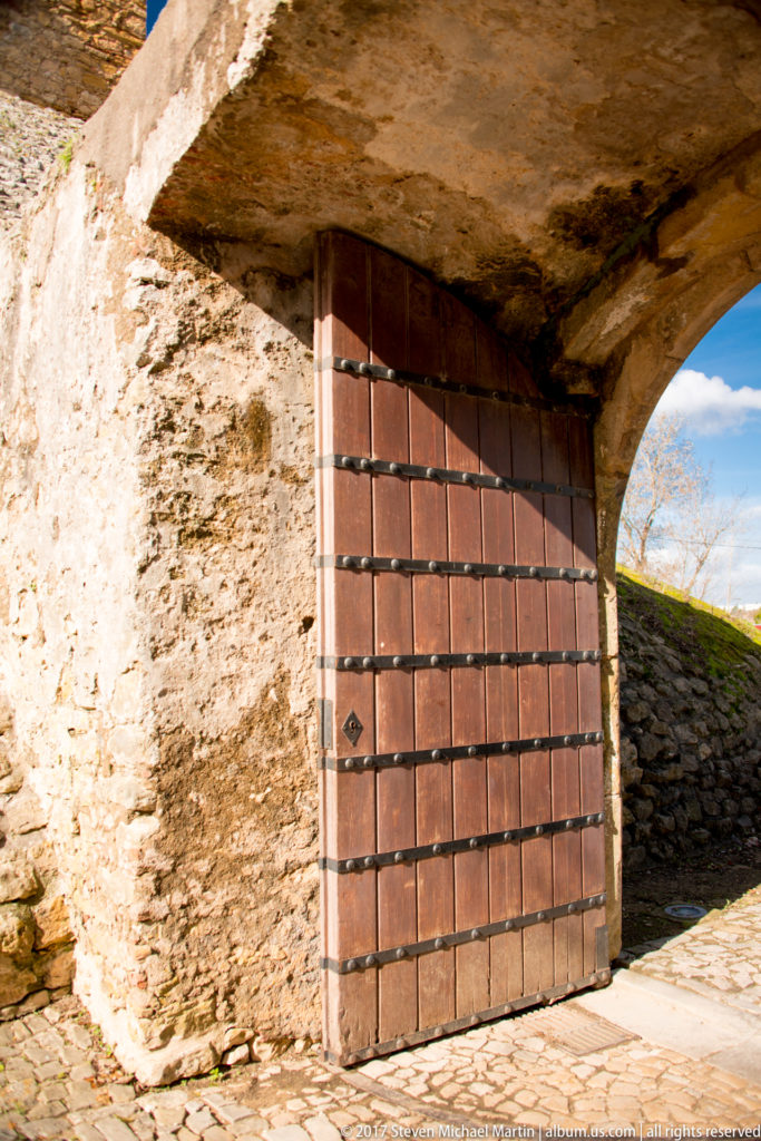 Large timber gate reinforced with dark iron straps and studs beneath a thick low stone passage, with daylight entering at right.