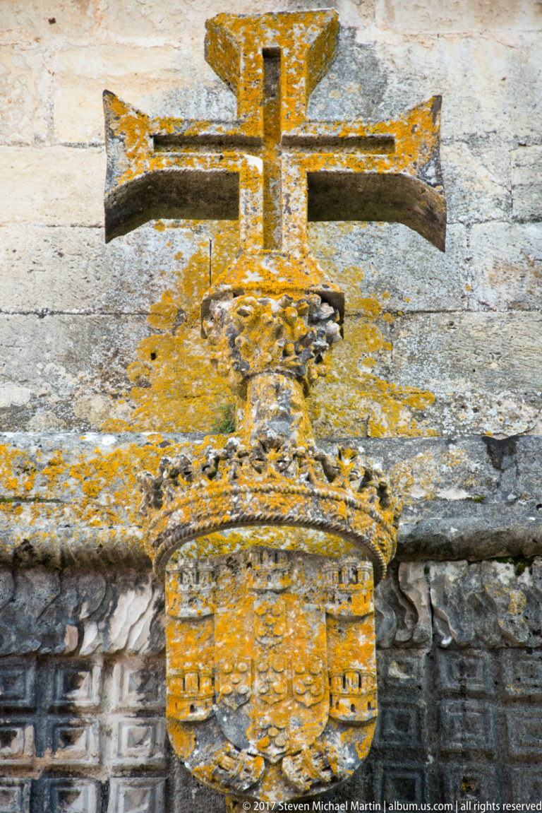 Large carved cross above an ornate heraldic shield on a weathered stone wall covered in patches of orange-yellow lichen.