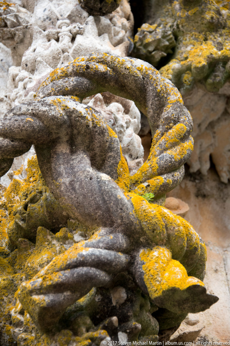 Close view of a weathered stone grotesque with looping carved forms and yellow lichen embedded in exterior ornament.