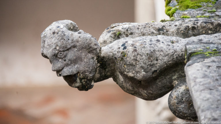 Weathered crouching stone gargoyle projecting horizontally from a wall, with softened facial features and patches of moss or lichen.