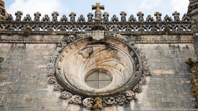Circular window in a dark stone wall surrounded by elaborate carved moldings, heraldic ornament, finials, and a cross above.