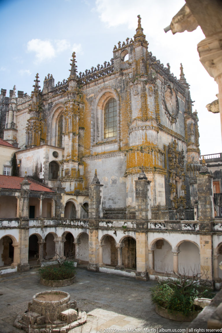 Wide courtyard view of the Convent of Christ church with ornate buttresses, pinnacles, arched windows, a circular opening, and lower arcades.