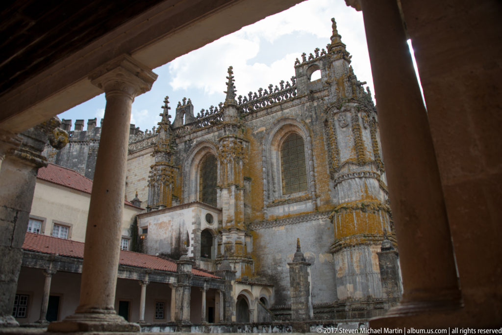 Ornate stone church exterior framed by cloister columns, with pinnacles, traceried windows, buttresses, and red-roofed lower ranges.