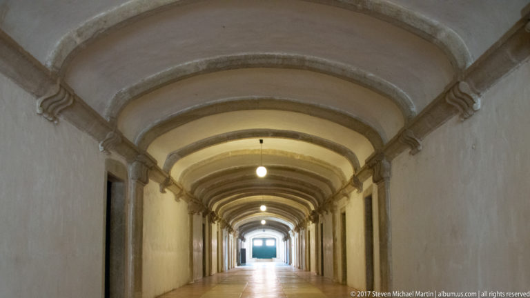 Long white corridor with repeated shallow arches, stone paving, doorways on both sides, and a bright opening at the far end.
