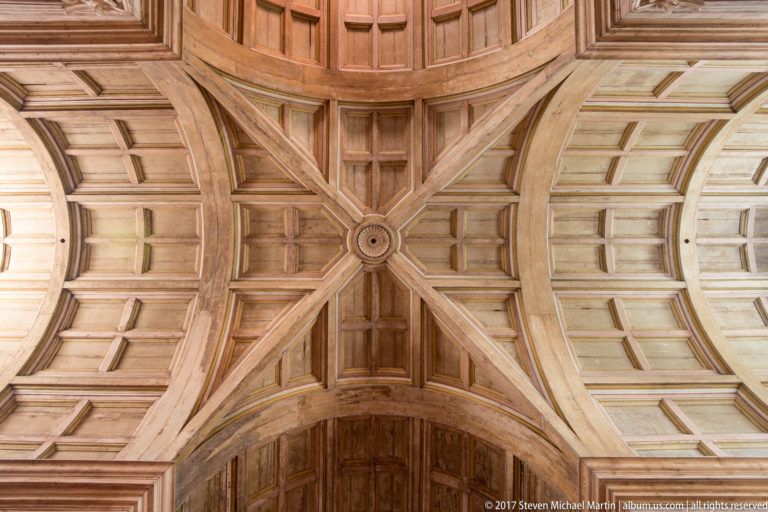 Close-up of intersecting wooden ceiling ribs forming an X around a circular boss amid curved coffered panels.