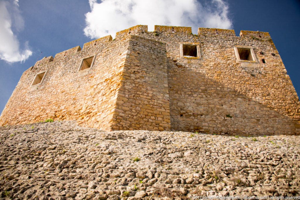Massive rectangular stone castle tower with deep openings rising above a rough sloped outer defensive wall.