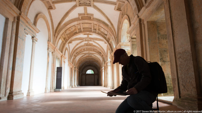 Long vaulted cloister corridor with repeating arches and a seated visitor silhouetted in the foreground.