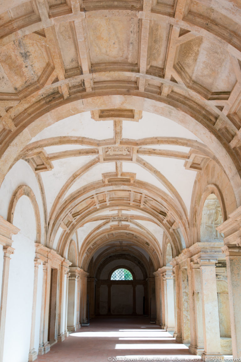 Long pale corridor beneath a coffered barrel vault, with repeated side arches leading toward a dark end wall.