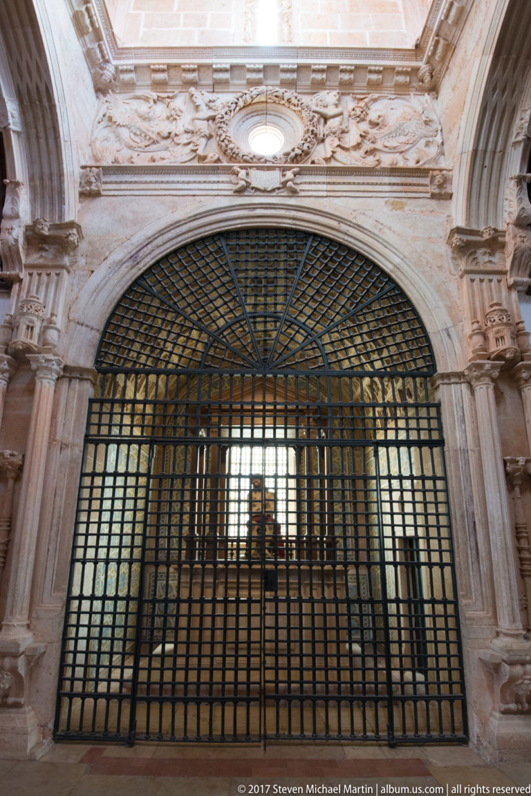 Large black iron grille with a fan-shaped arched top set inside an ornate pale stone portal.